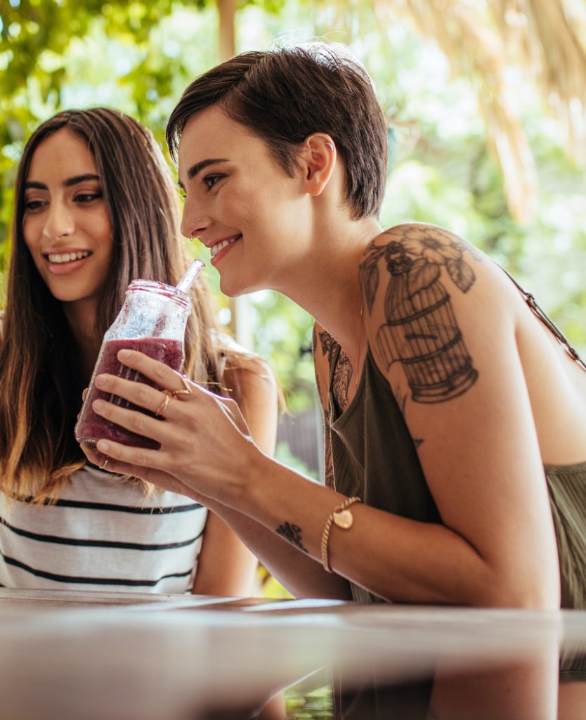 Photo of a woman with tattoos on her arm drinking a smoothie next to a friend